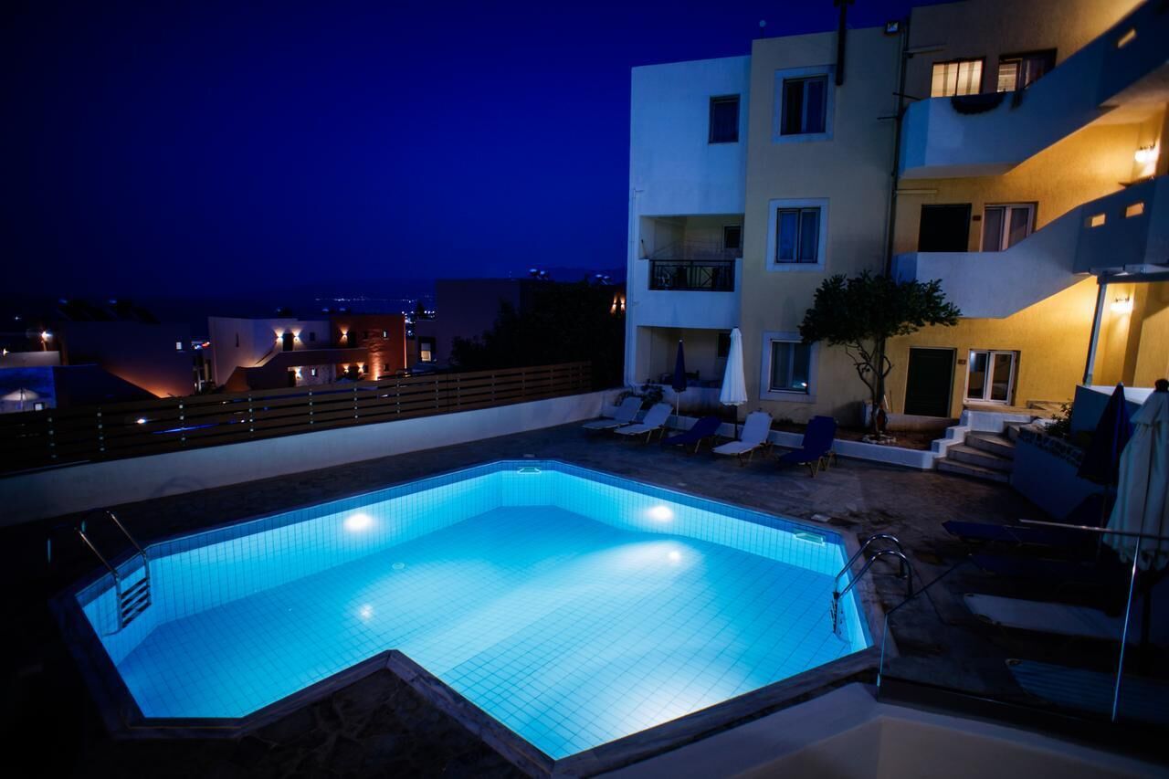 A rectangular swimming pool with a tiled deck, surrounded by lounge chairs and umbrellas, adjacent to a multi-story building with balconies and outdoor lighting.