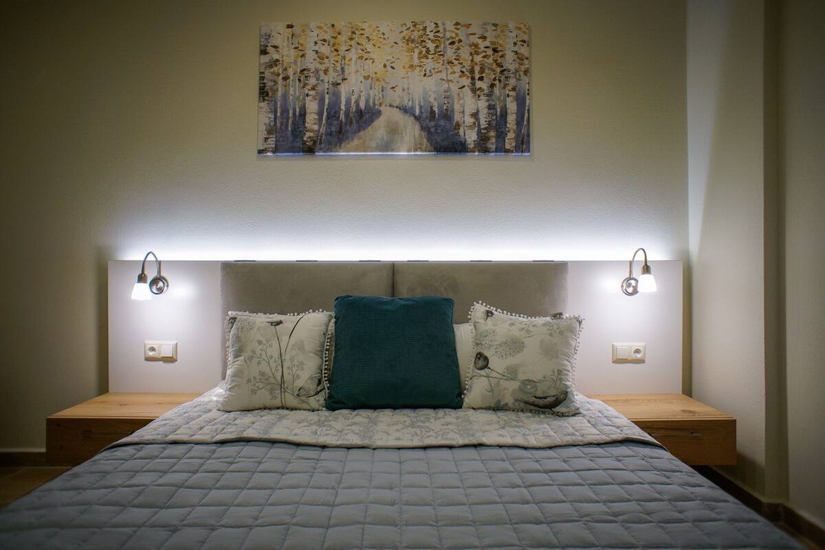 A bedroom with a bed, two bedside tables, a headboard with integrated lighting, and a wall-mounted painting.