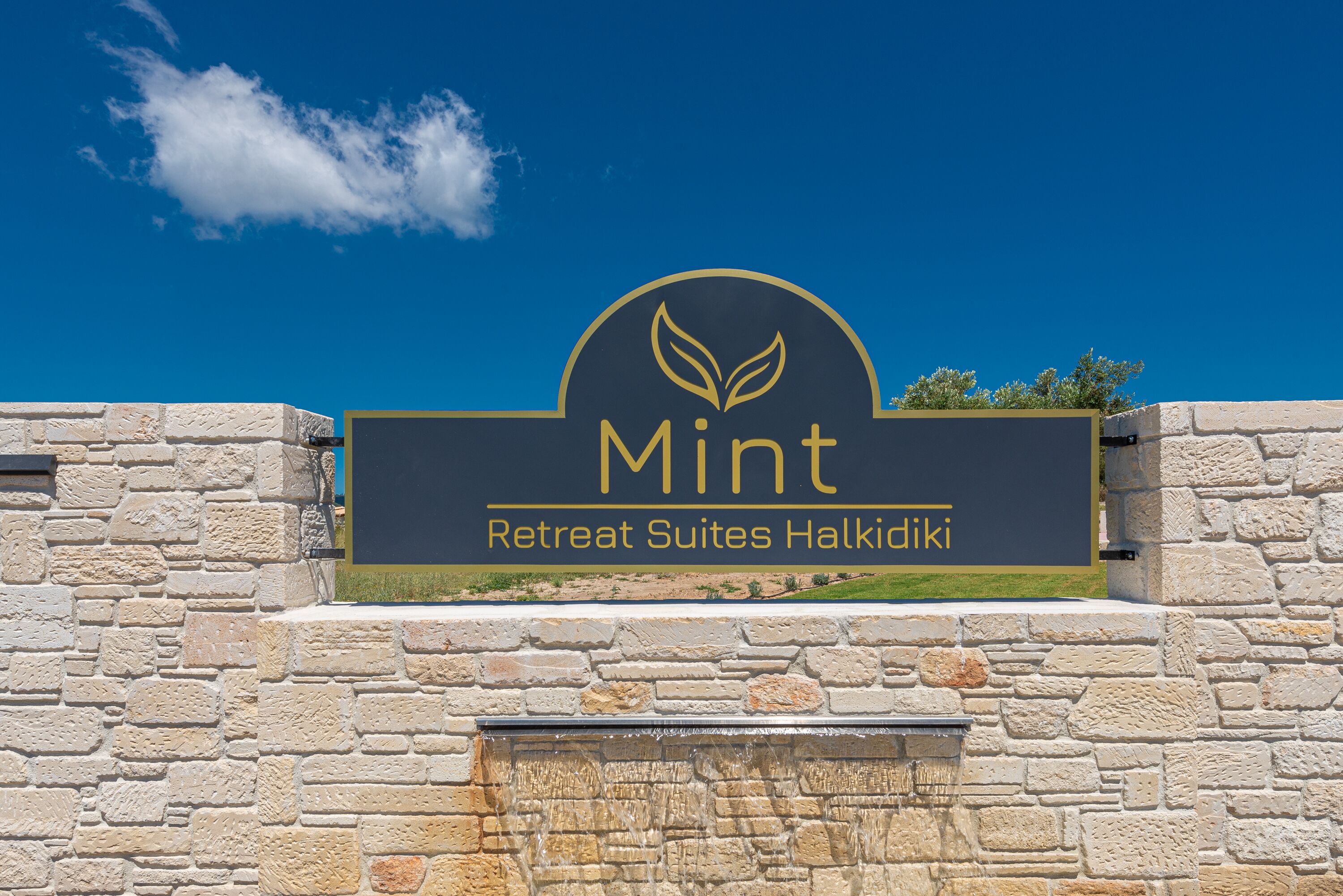 Un cartel para 'Mint Retreat Suites Halkidiki' con un muro de piedra y un cielo azul despejado.