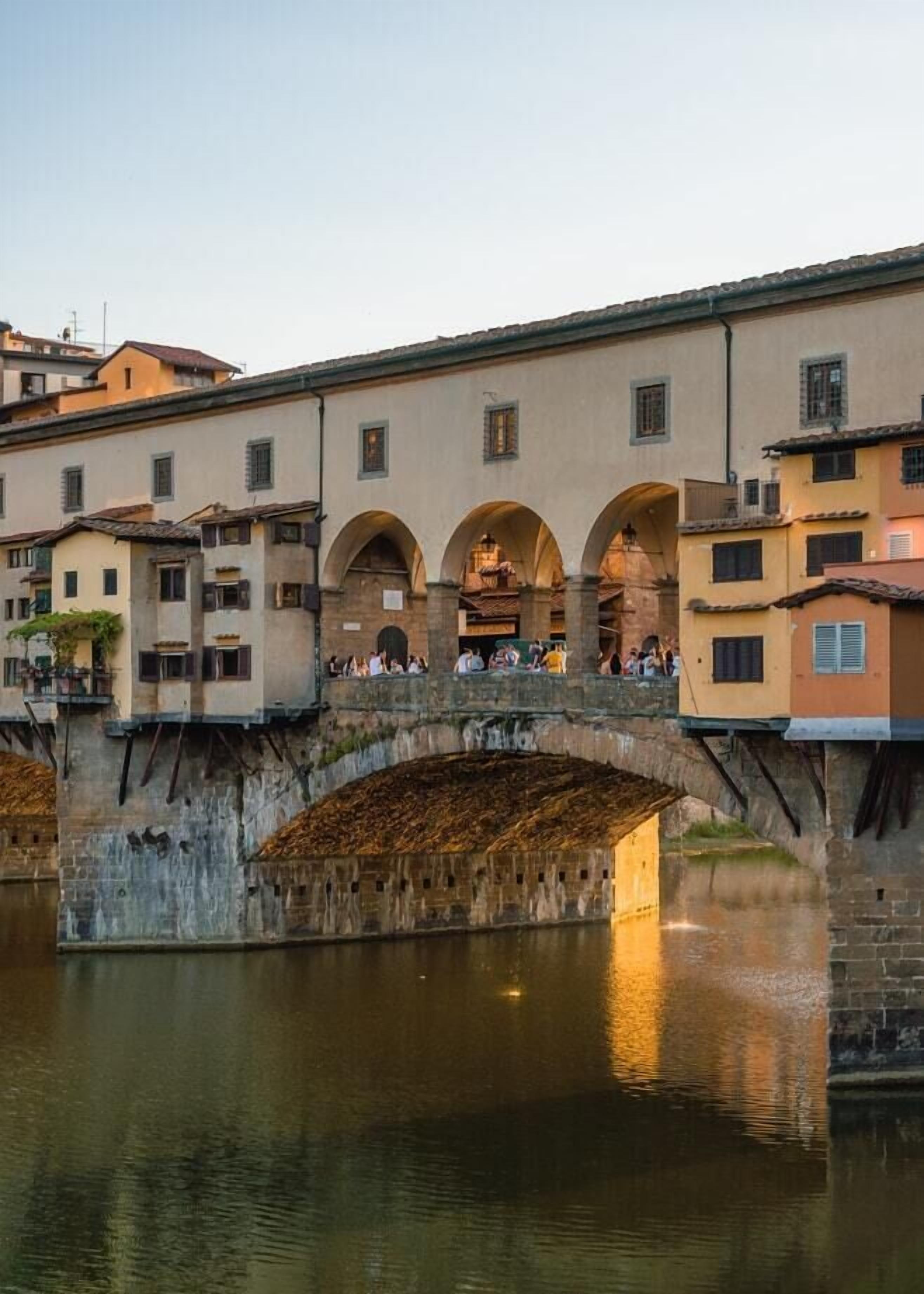 A stone bridge with arches spanning a calm river, surrounded by historic buildings with multiple windows and balconies.