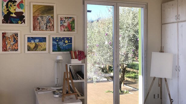 A room with a white table, a lamp, and a painting. There is a sliding glass door leading to an outdoor area with a tree and a fence. - SUITE LA GINESTRA among historic olive trees (Eboli SALERNO)