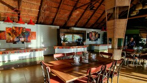 A restaurant with a rustic interior, featuring a thatched roof, wooden beams, and a white brick wall. There are red pendant lights, a buffet counter, and several dining tables with chairs. - The Wallow Lodge (Victoria Falls)
