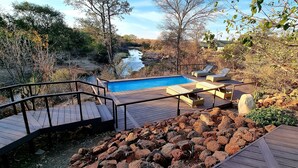 A wooden deck with a pool, lounge chairs, and a view of a river. - The Wallow Lodge (Victoria Falls)