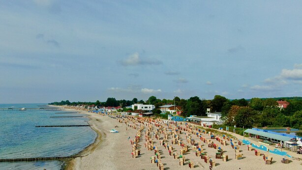 A beach with rows of wooden beach chairs and umbrellas, a pier extending into the water, and buildings along the shore. - Störtebeker 4 (Kellenhusen)