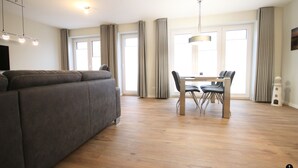A modern living room with a grey sofa, a dining table with chairs, and large windows with curtains. - Störtebeker 4 (Kellenhusen)