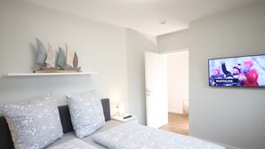A bedroom with a bed, pillows, a nightstand, a TV displaying a marathon event, and a shelf with decorative items. - Störtebeker 4 (Kellenhusen)