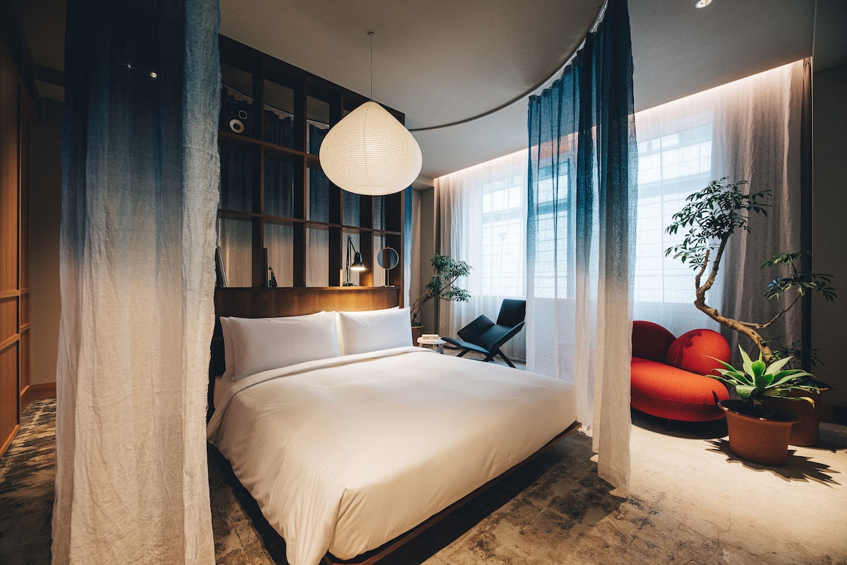 A modern bedroom with a large bed, a reading chair, a small table, and a potted plant.