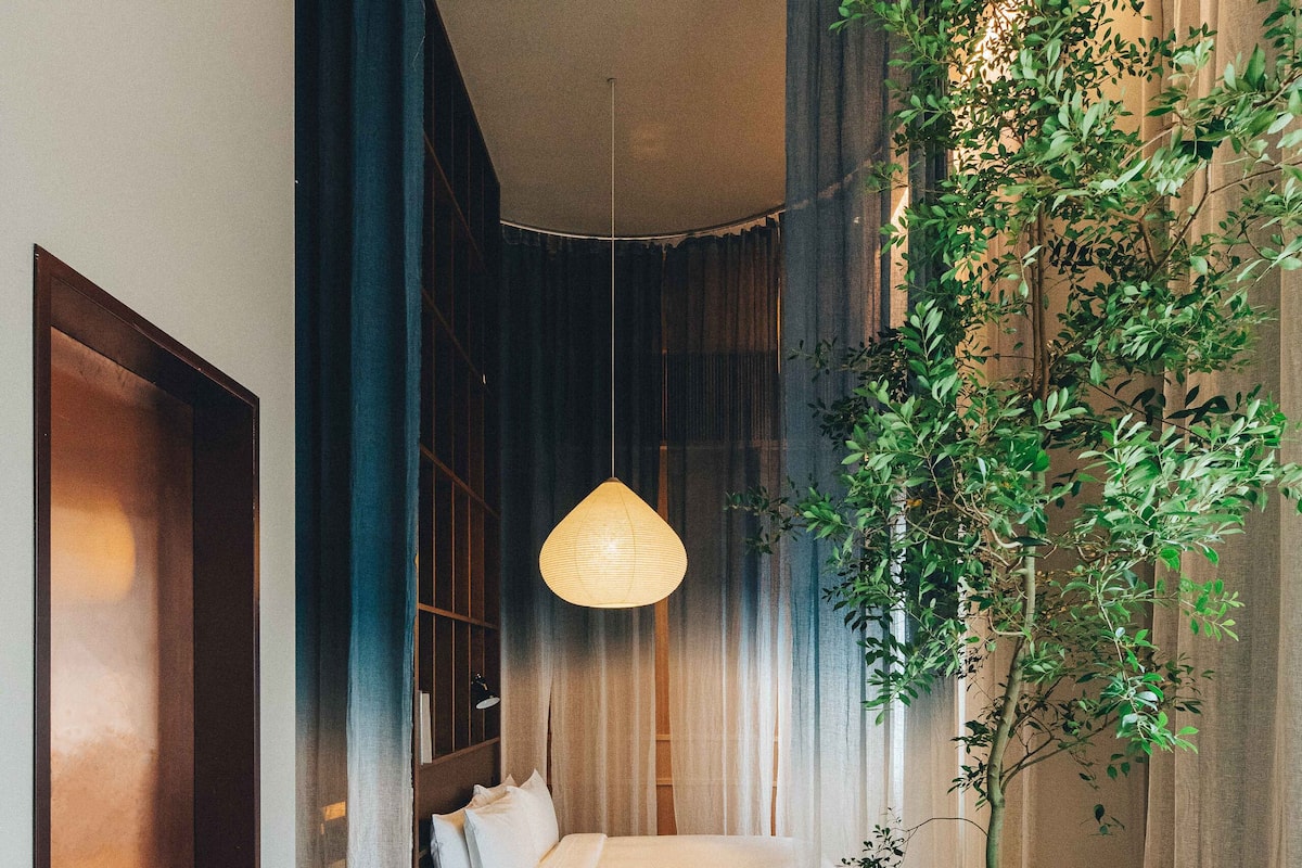 A hotel room with a bed, a large potted plant, a hanging lamp, and a red chair.