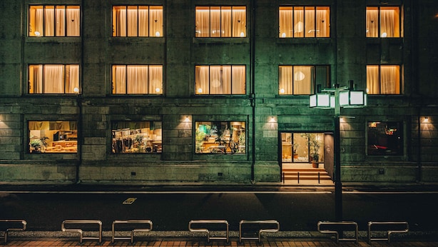 Un edificio de varios pisos con ventanas iluminadas, una entrada visible y mesas al aire libre.