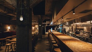 Una zona de bar con taburetes de madera, una barra larga y una cocina al fondo.