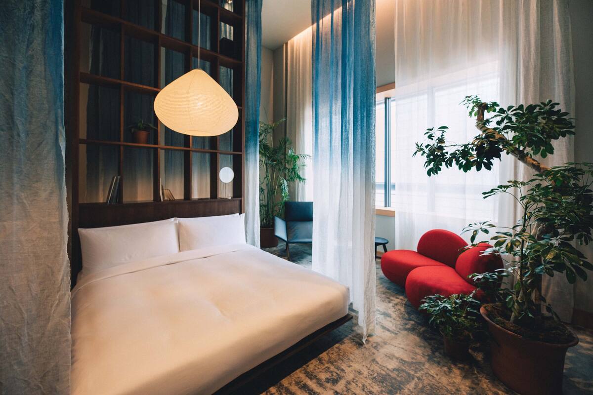 A hotel room with a large bed, a hanging light fixture, a potted plant, and a view of the outdoors through sheer curtains.