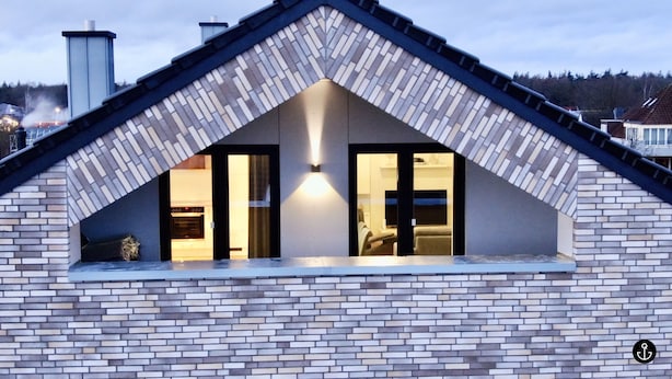 Uma casa moderna com fachada de tijolos, telhado inclinado e duas janelas visíveis com iluminação interna.