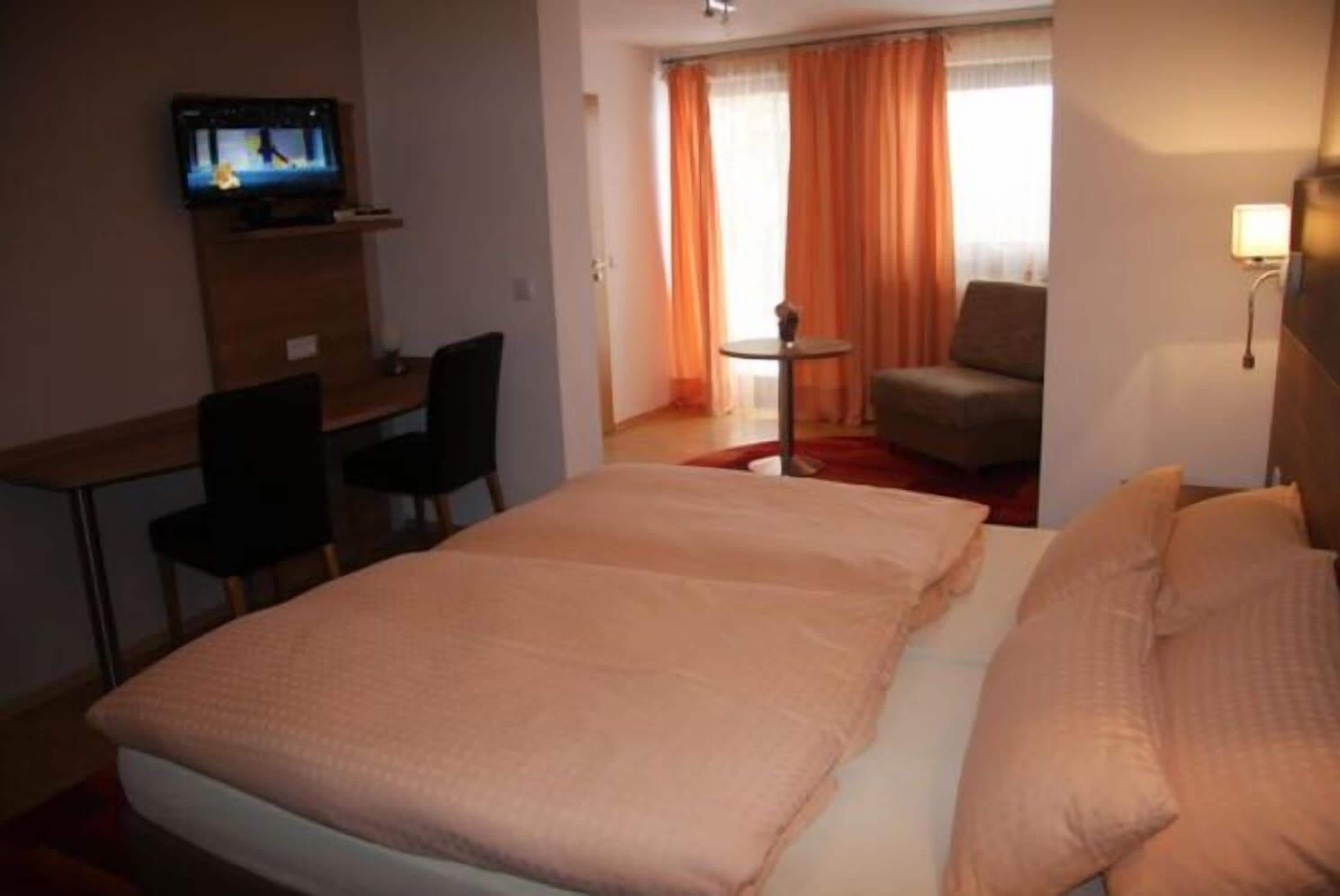 Una habitación de hotel con una cama, un escritorio con una silla, un televisor, una mesita con una silla y una ventana con cortinas.