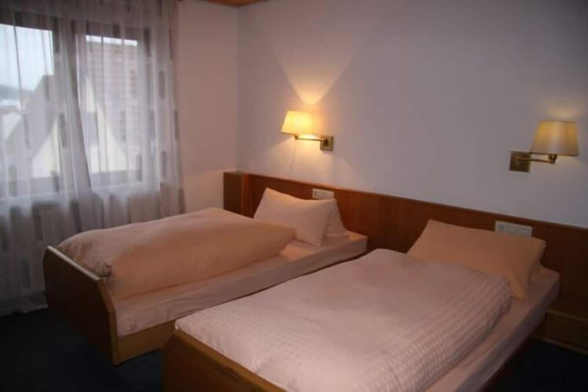 Habitación de hotel con dos camas individuales, una ventana con cortinas traslúcidas y lámparas de pared.