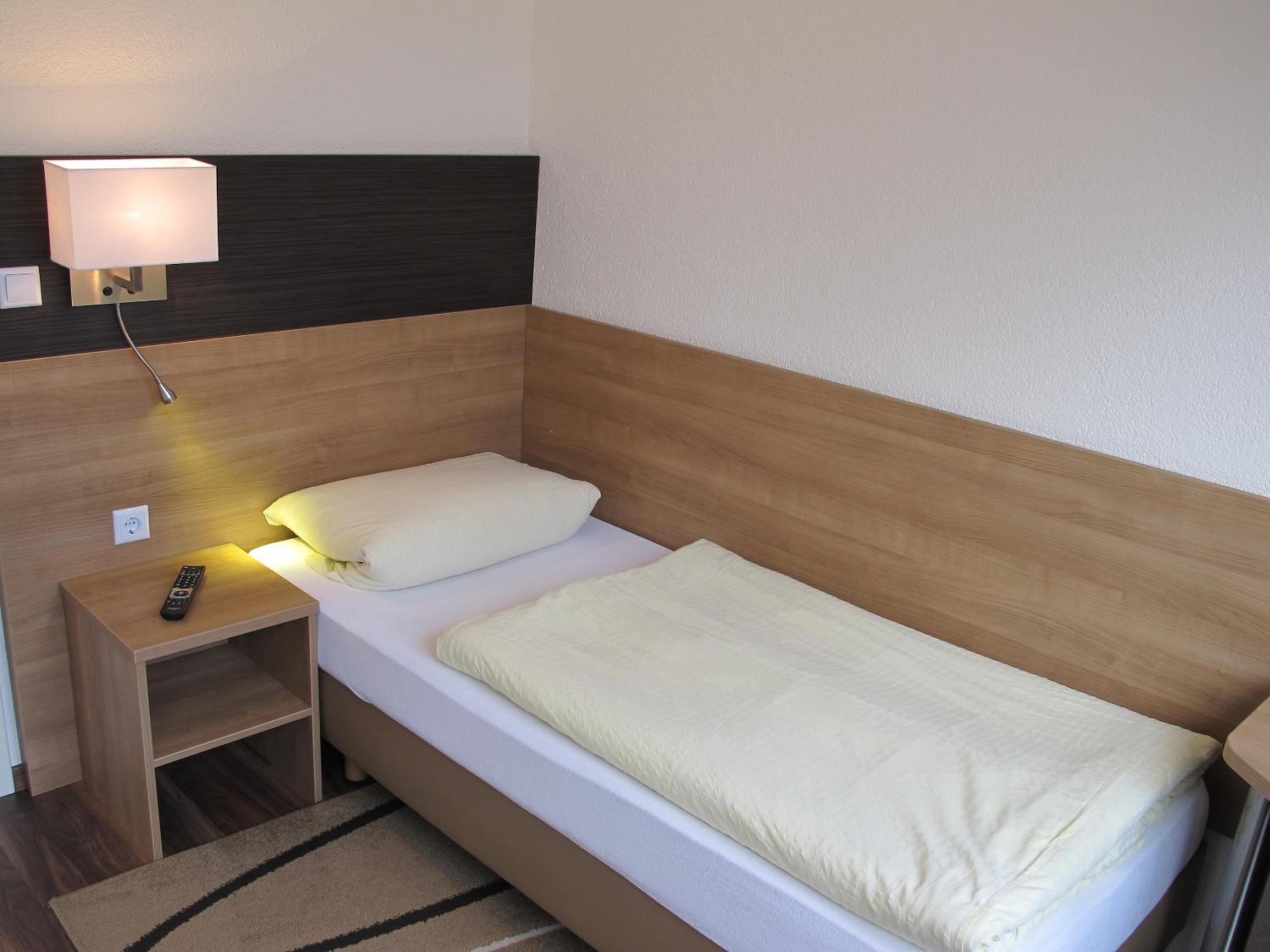 A single bed with a wooden headboard, a bedside table with a remote control, and a wall-mounted lamp.