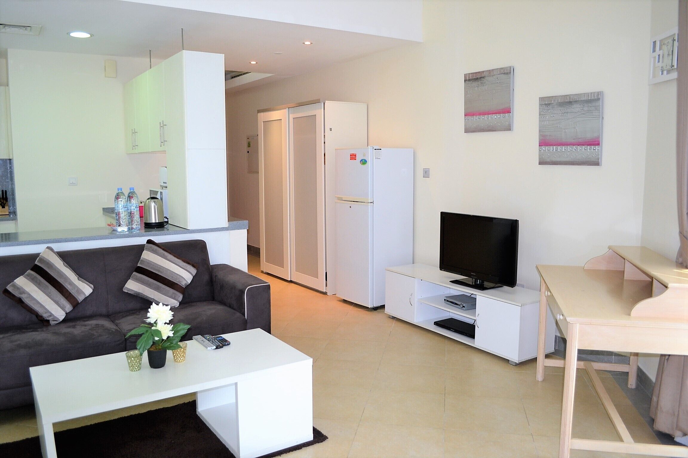 Uma sala de estar moderna com sofá, mesa de centro, televisão e uma área de cozinha com armários e geladeira.