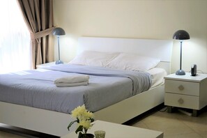 A neatly made bed with striped bedding, a bedside table with a lamp, and a nightstand with a phone. - Bright furnished studio near metro in Tecom - ALIA (Dubai)