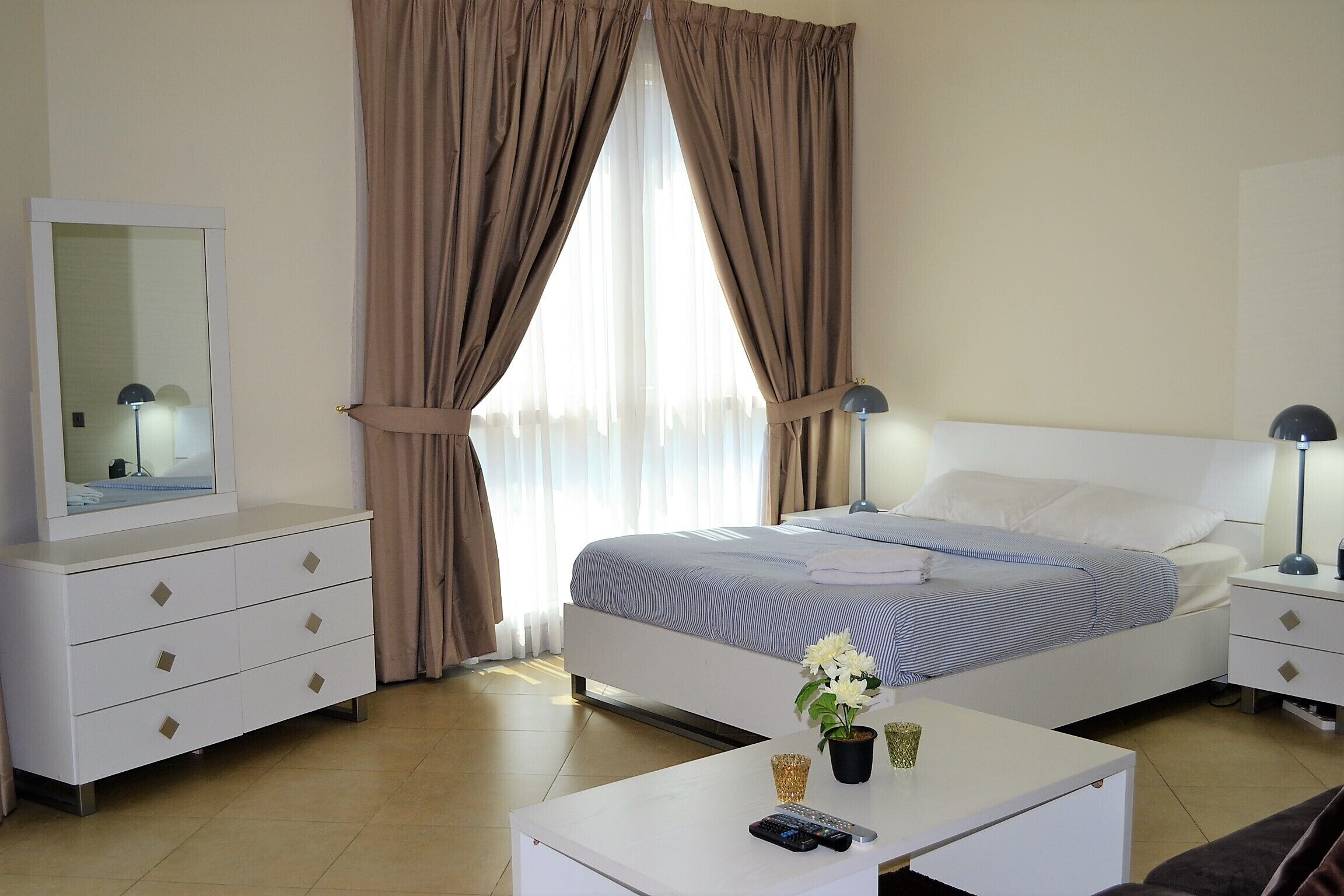 Habitación de hotel con cama, mesitas de noche, cómoda, espejo y ventana con cortinas.