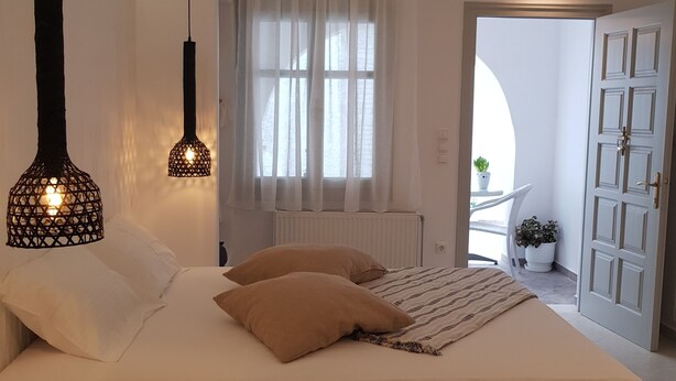 A modern bedroom with a large bed, two hanging pendant lights, a white radiator, and a view of the outside through a window with white curtains. - Exclusive Studios Naxos Town (Naxos)