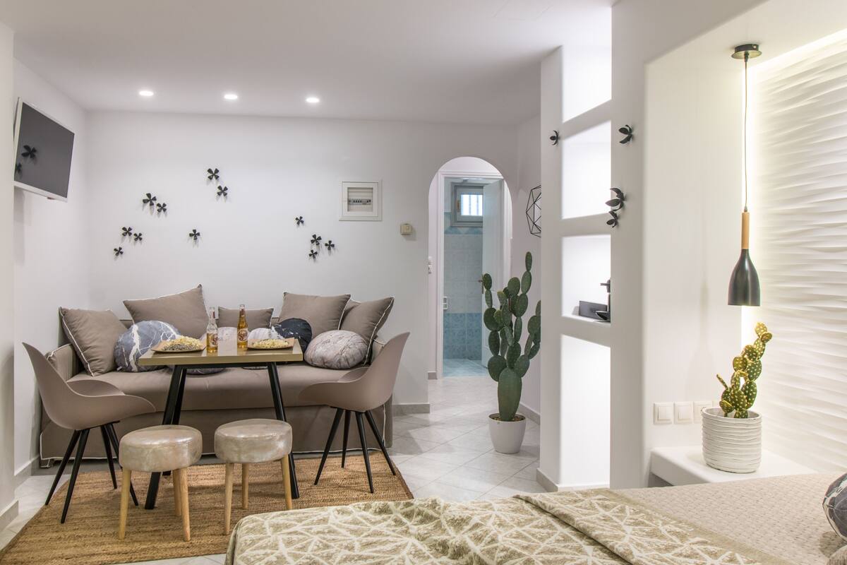 A modern living room with a sofa, coffee table, and two ottomans. There is a cactus plant and a wall-mounted TV.