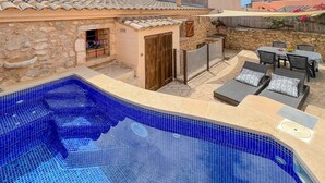 Una piscina amb un videt de rajola, un edifici de pedra amb una porta de fusta, i mobiliari exterior amb coixins.