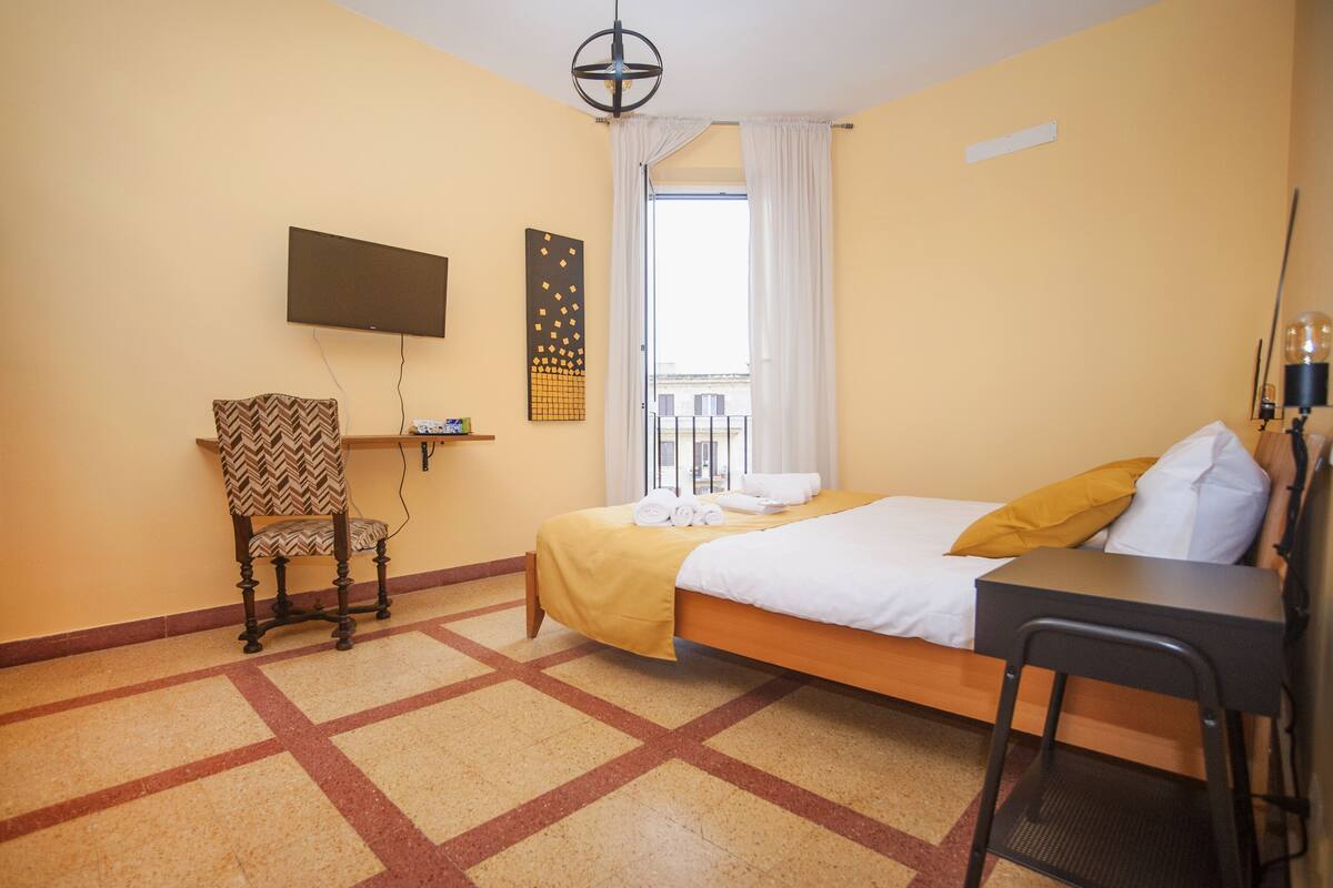 A hotel room with a bed, a desk, a chair, a TV mounted on the wall, and a window with curtains.