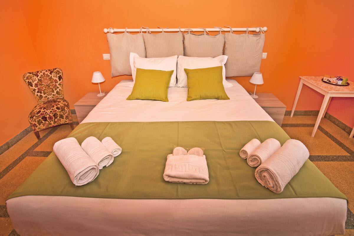 A hotel room with a bed, green bedspread, two pillows, a chair, two lamps, a small table with a tray, and rolled towels on the bed.
