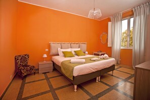 A bedroom with an orange wall, a bed with green and white bedding, a patterned armchair, a bedside table with a lamp, and a window with curtains. - Martin (Rome)
