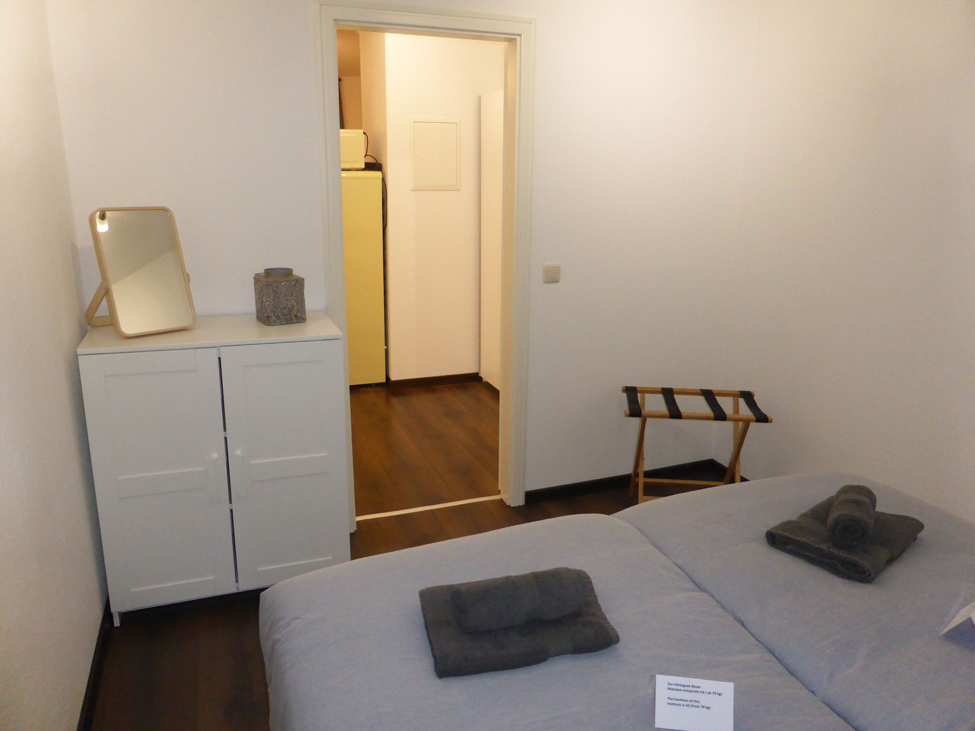 A hotel room with a bed, a bedside table with a lamp, a mirror, and a small wooden chair.