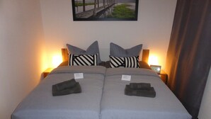 A double bed with grey bedding, two pillows, two folded towels, and a wooden frame. There are two bedside tables with lamps, a framed picture on the wall, and a clock on the wall. - Apartment Goethe (Jena)
