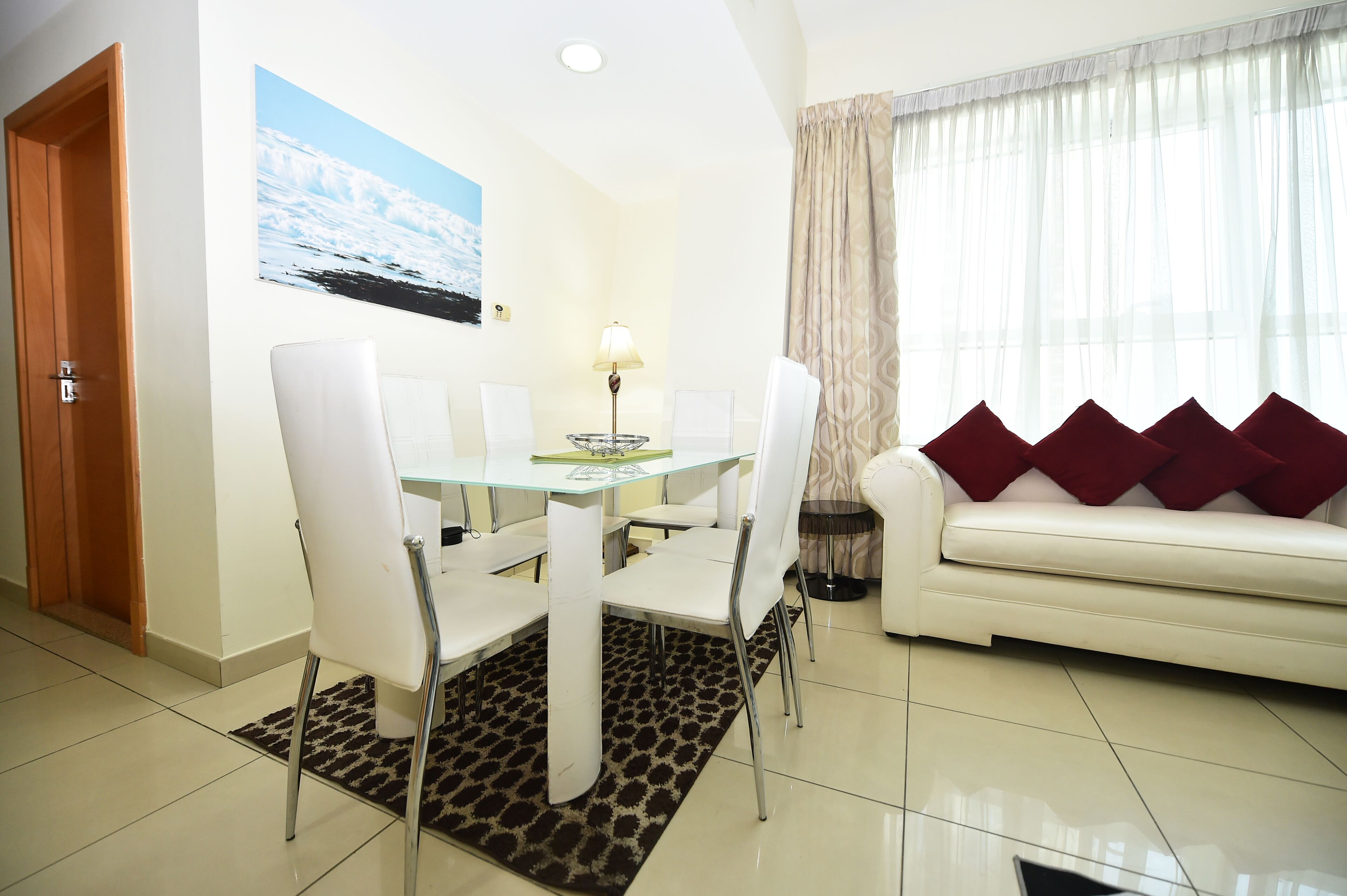 A dining area with a glass table and white chairs, a white sofa with red cushions, and a painting of a snowy landscape on the wall.