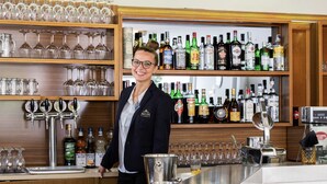 A bar area with shelves of liquor bottles, a bartender, and a bar counter with glasses and a cash register. - Miga Hotel Grado Centro (Grado)