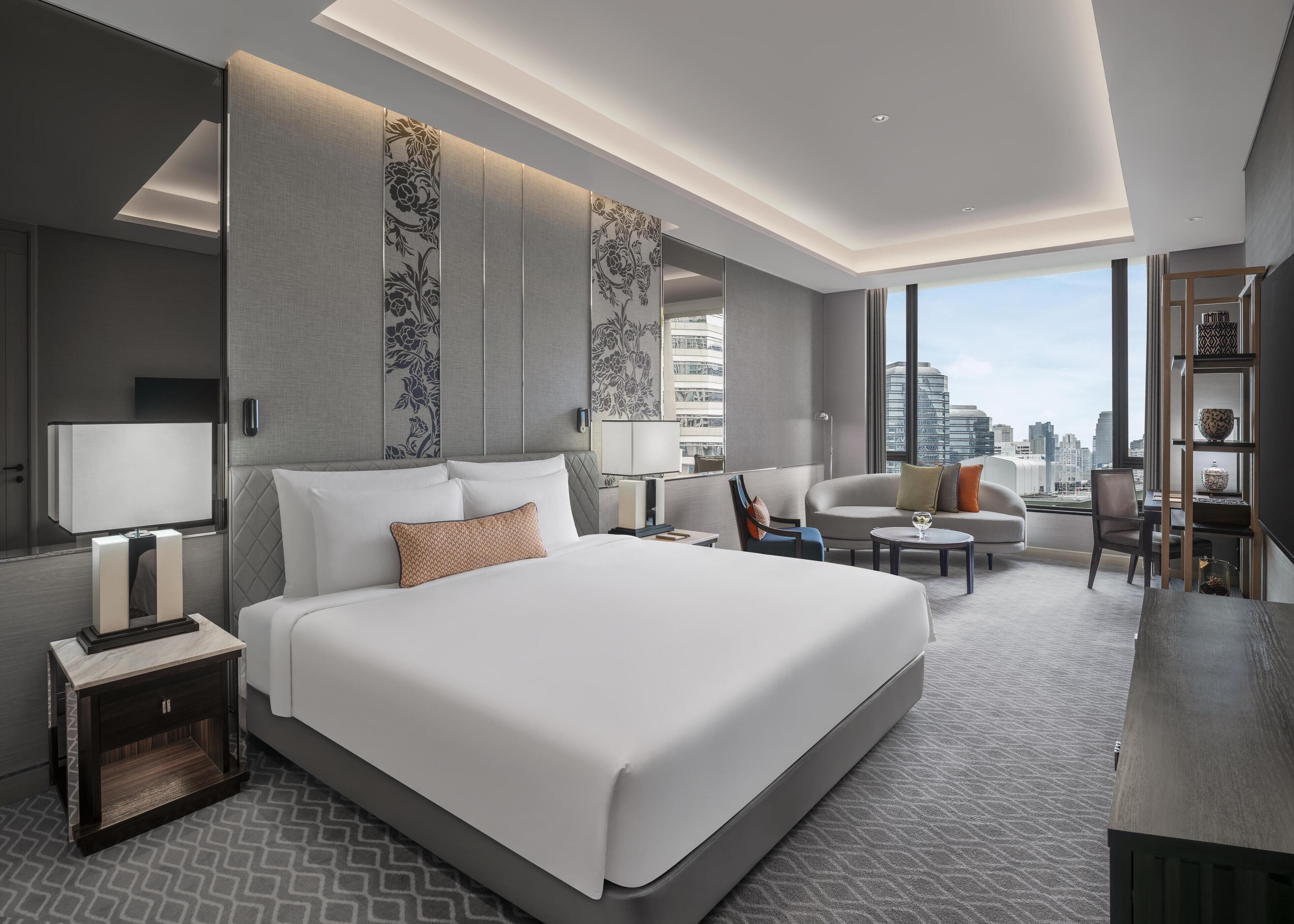 A modern hotel room with a large bed, a sofa, a chair, a small table, and a bookshelf. The room has large windows with a city view.