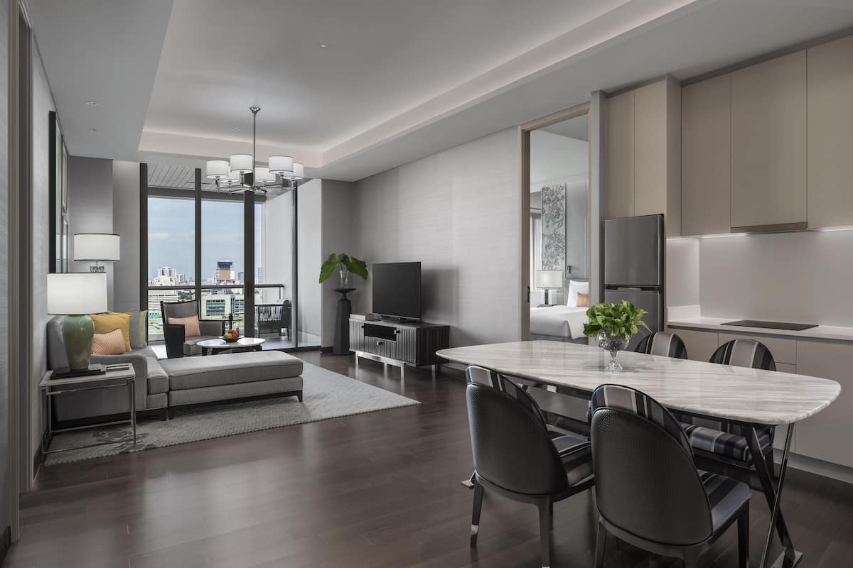 Una sala de estar moderna con sofá, mesa de centro y televisor. Hay un comedor con mesa y sillas. La habitación tiene un ventanal con vistas a la ciudad.