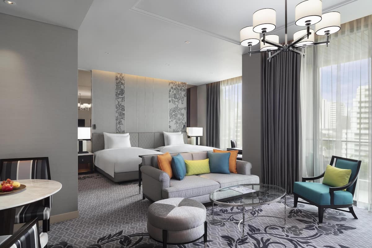 Una habitación de hotel moderna con una cama grande, un sofá, una silla, una mesa de comedor con fruta y vistas a la ciudad.