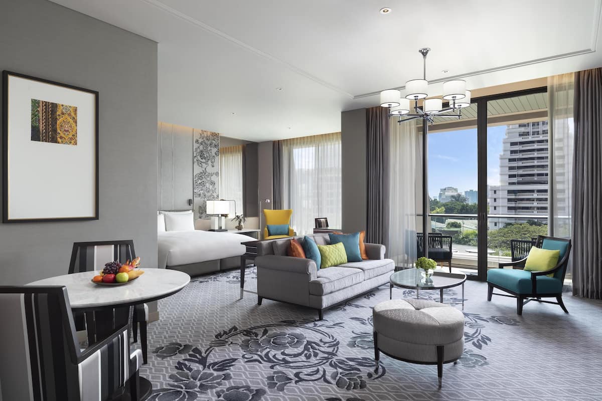 Habitación de hotel moderna con una cama grande, un sofá gris, una mesa de comedor con fruta y vistas a la ciudad a través de amplias ventanas.