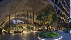Un edificio moderno con fachada de vidrio curvo, un atrio central y una zona ajardinada con un árbol.