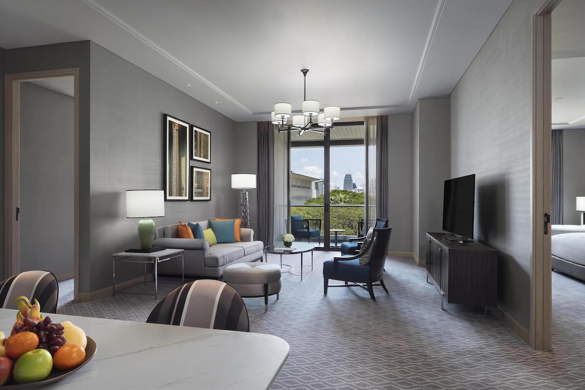 Una habitación de hotel moderna con sofá, sillón, mesa de comedor con fruta, televisor y balcón con vista a la ciudad.