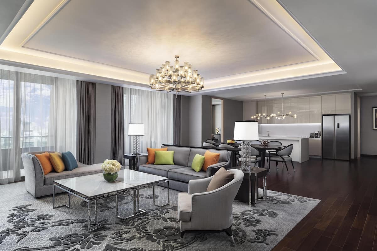Una sala moderna con sofá, sillón y mesa de centro, una araña y una zona de cocina al fondo.