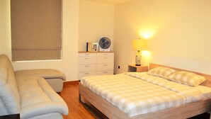A bedroom with a bed, a sofa, a bedside table with a lamp, a fan, and a window with blinds. - Stylish 1BR near MOE - MRD (Dubai)