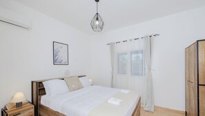 A bedroom with a wooden bed, white bedding, a bedside table with a lamp, a hanging light fixture, and a window with curtains. - Beachfront Cabana @ EcoBeach Hotel (Gennadi)