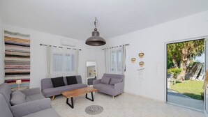 A modern living room with a sofa, armchair, and coffee table. There is a large window with curtains, a hanging light fixture, and a view of a garden. - Beachfront Cabana @ EcoBeach Hotel (Gennadi)