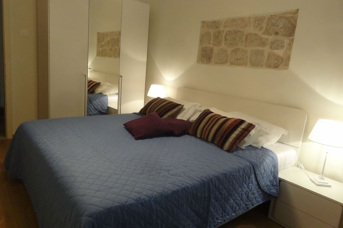 A bedroom with a large bed, a wardrobe, a nightstand with a lamp, and a stone wall decoration.