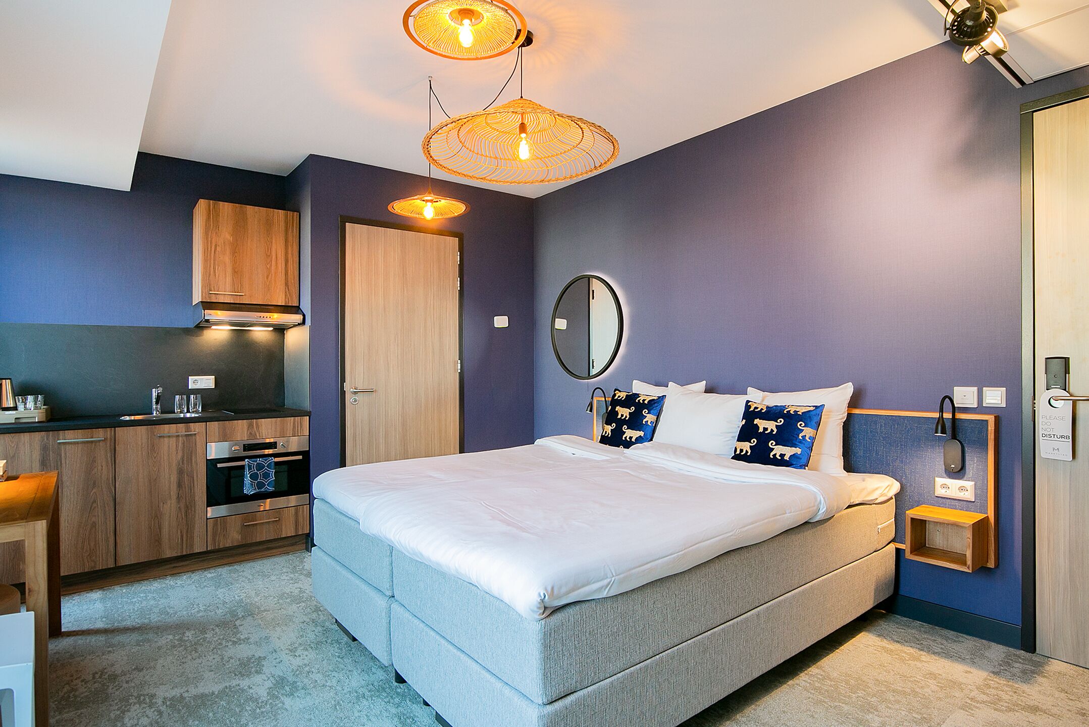 Un dormitorio moderno con una cama grande, mesitas de noche, un espejo y una cocina integrada con armarios y electrodomésticos.