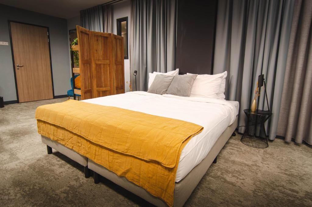 Een net opgemaakt bed met een gele deken, grijze gordijnen en een houten garderobe in een slaapkamer.