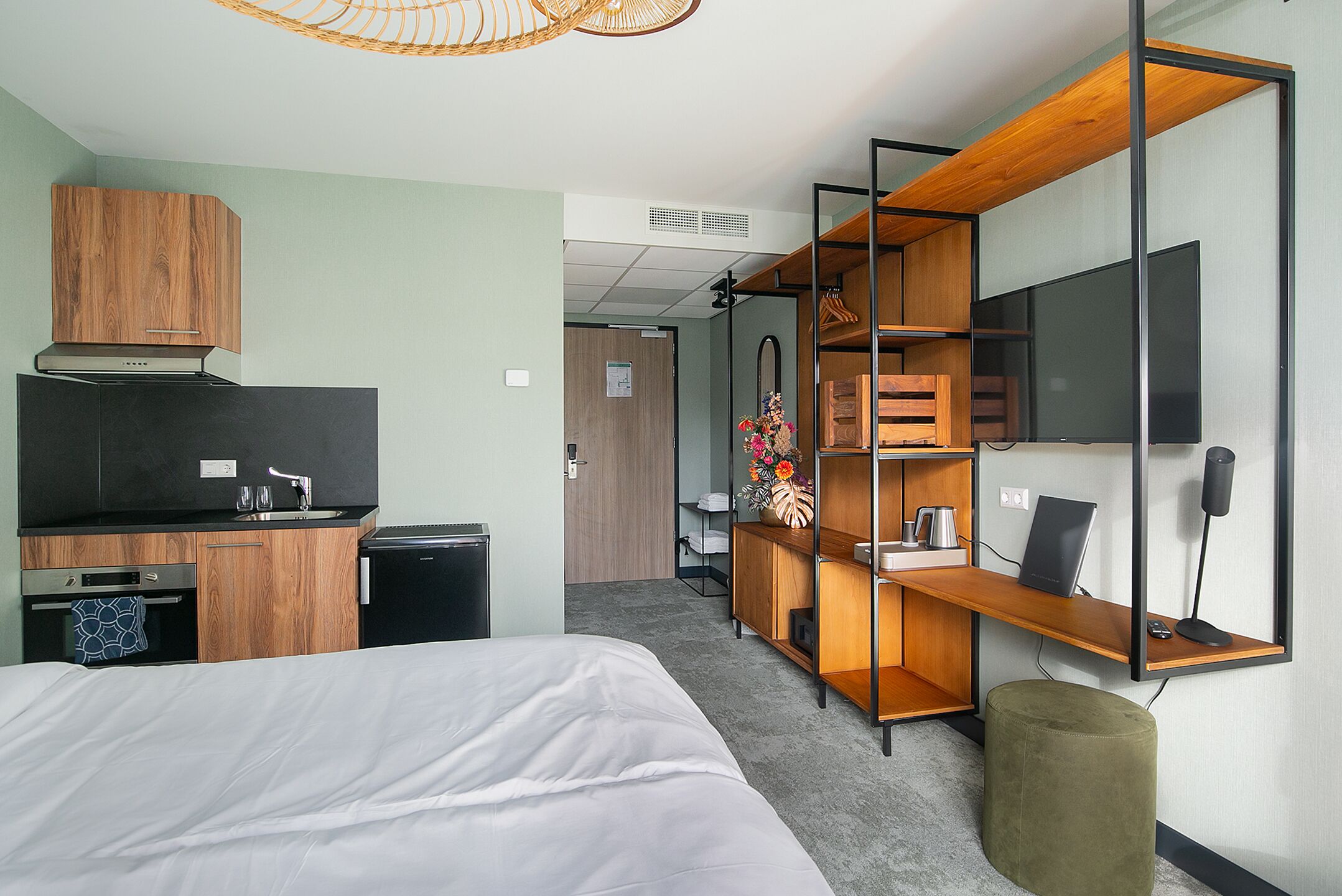 Ein modernes Hotelzimmer mit einer Küchenzeile, einem Bett, einem Schreibtisch mit Computer und einem großen Spiegel.