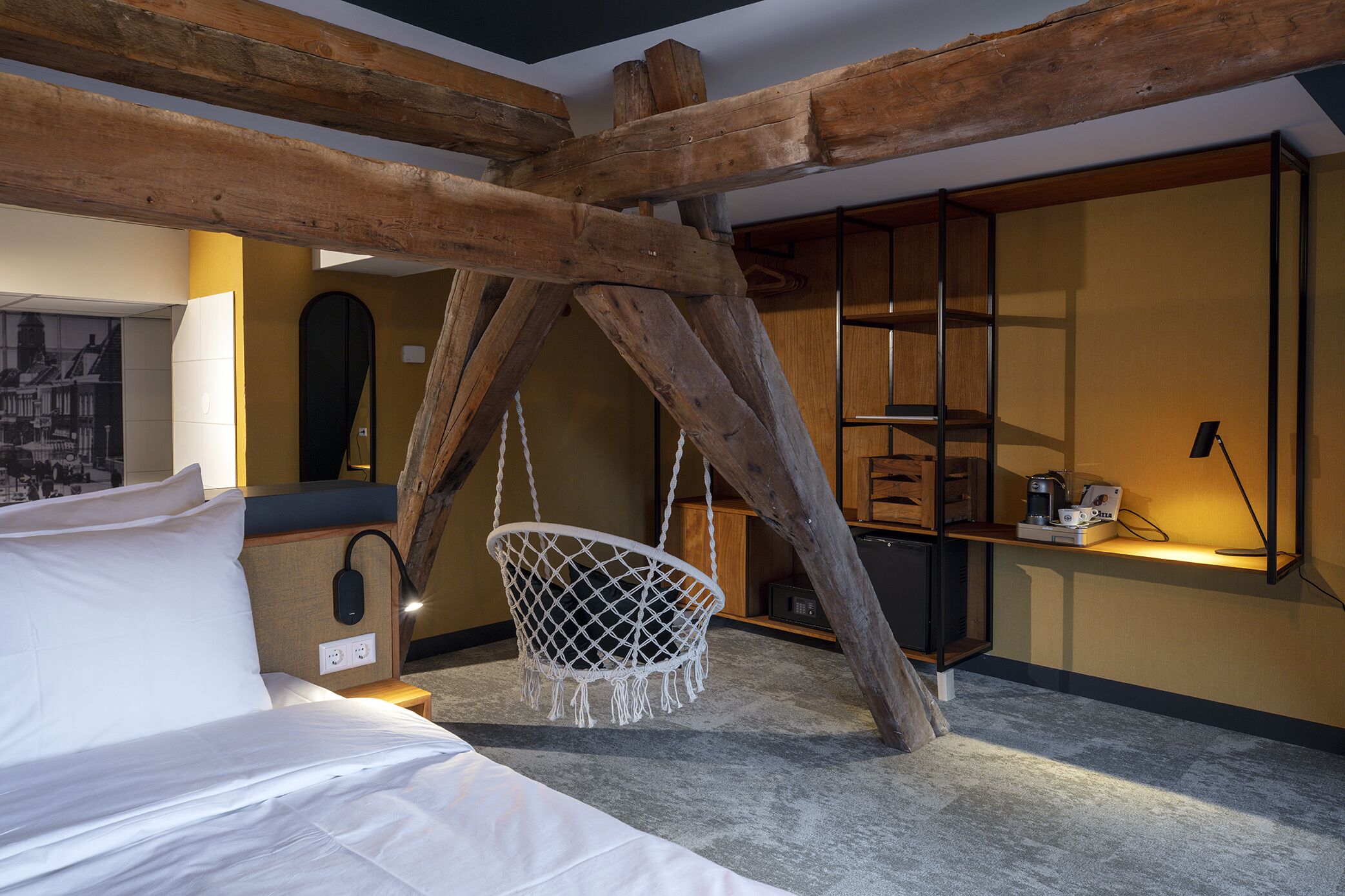 A room with a bed, a hanging chair, wooden beams, a shelf with items, and a wall-mounted lamp.