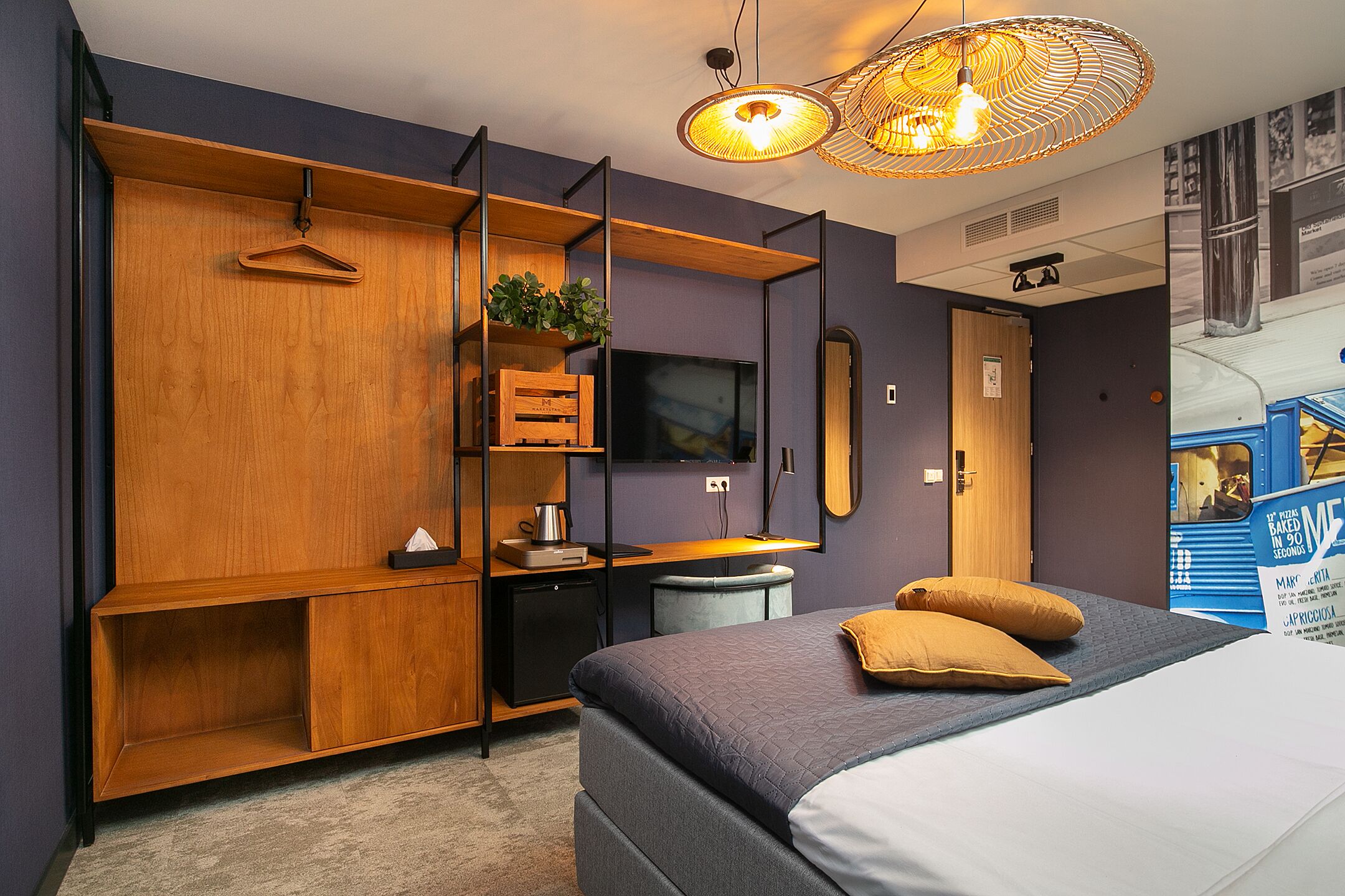 A modern hotel room with a bed, a wooden wardrobe, a desk with a computer, and a hanging light fixture.