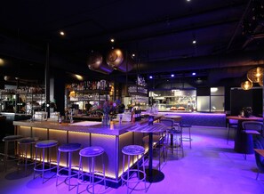 Una zona de bar con mostrador, taburetes y estanterías llenas de botellas y vasos.