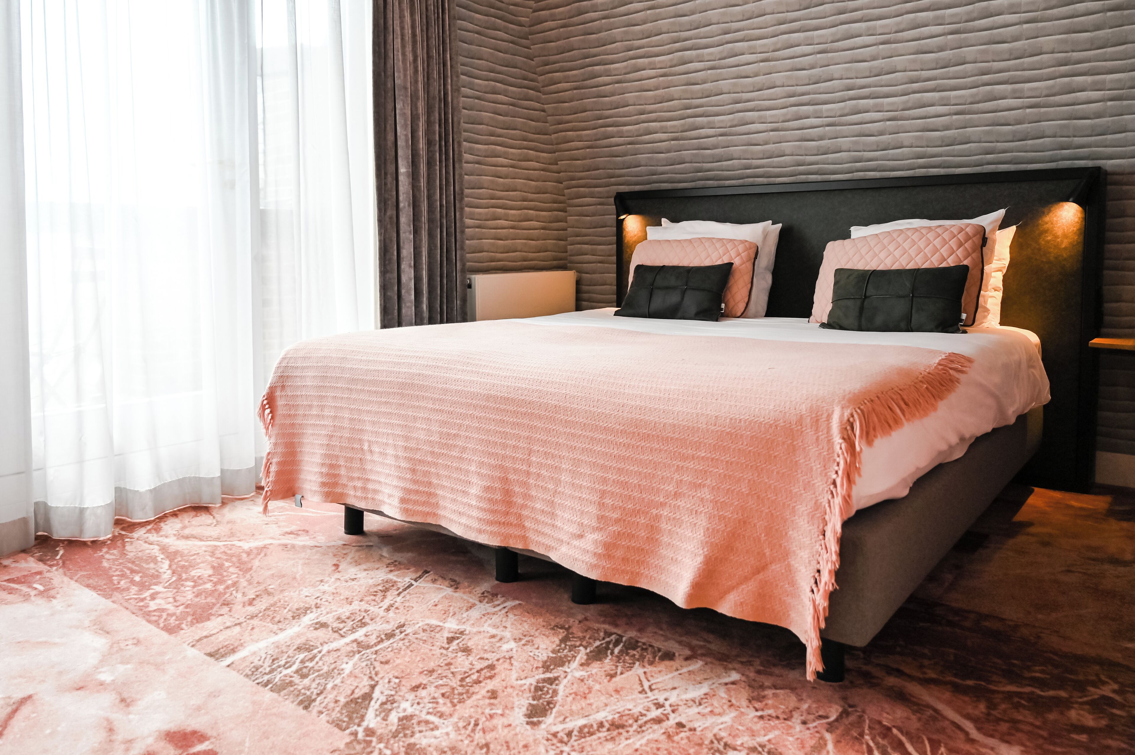 A neatly made bed with a pink bedspread and multiple pillows, a bedside table with a lamp, and a textured wall behind the bed.